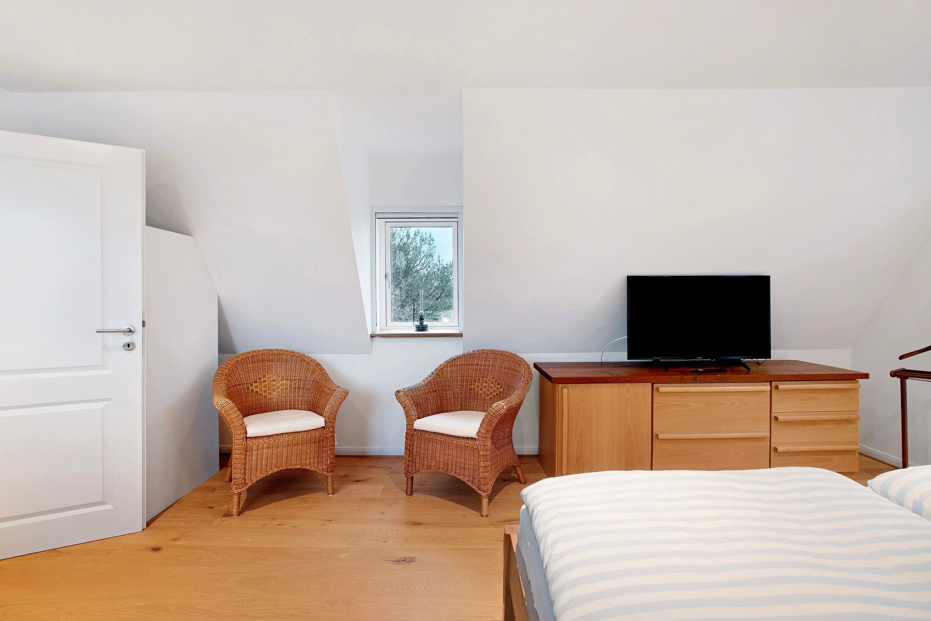 Een slaapkamer met een bed, twee rotan fauteuils, een houten dressoir, een televisie en een raam.