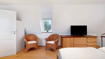 Een slaapkamer met een bed, twee rotan fauteuils, een houten dressoir, een televisie en een raam.