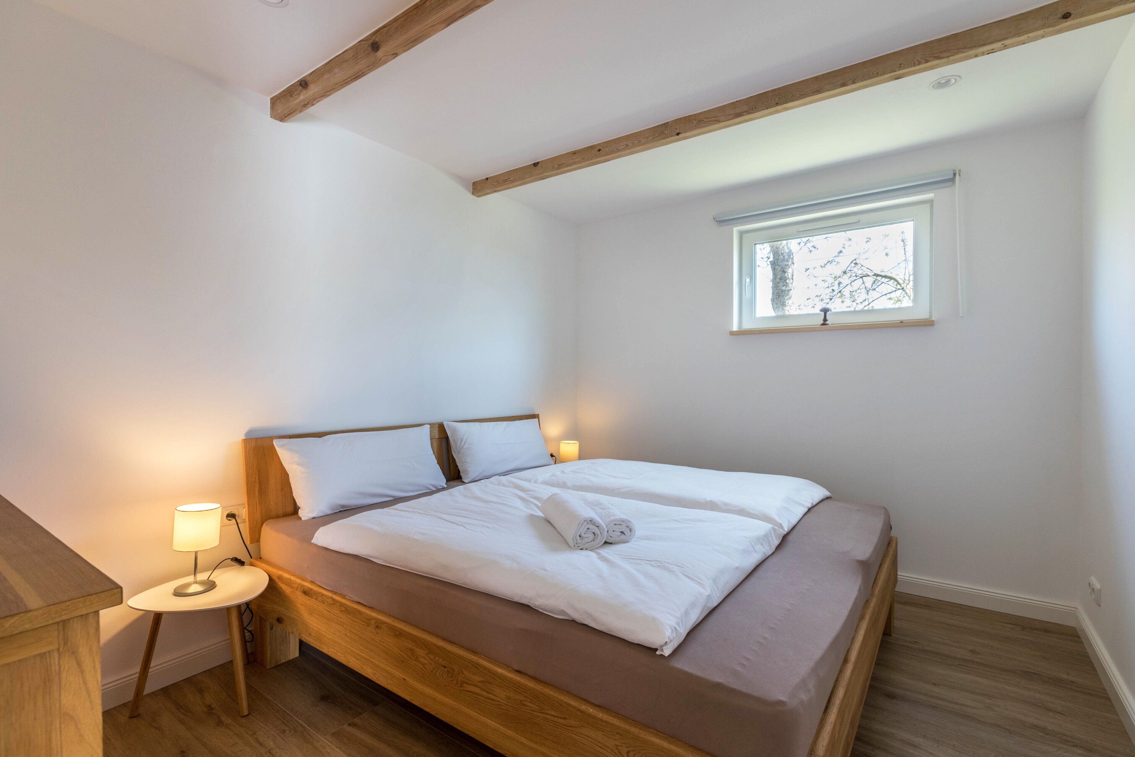 Un dormitorio con una cama de madera, una mesita de noche con lámpara y una ventana con vistas a árboles.