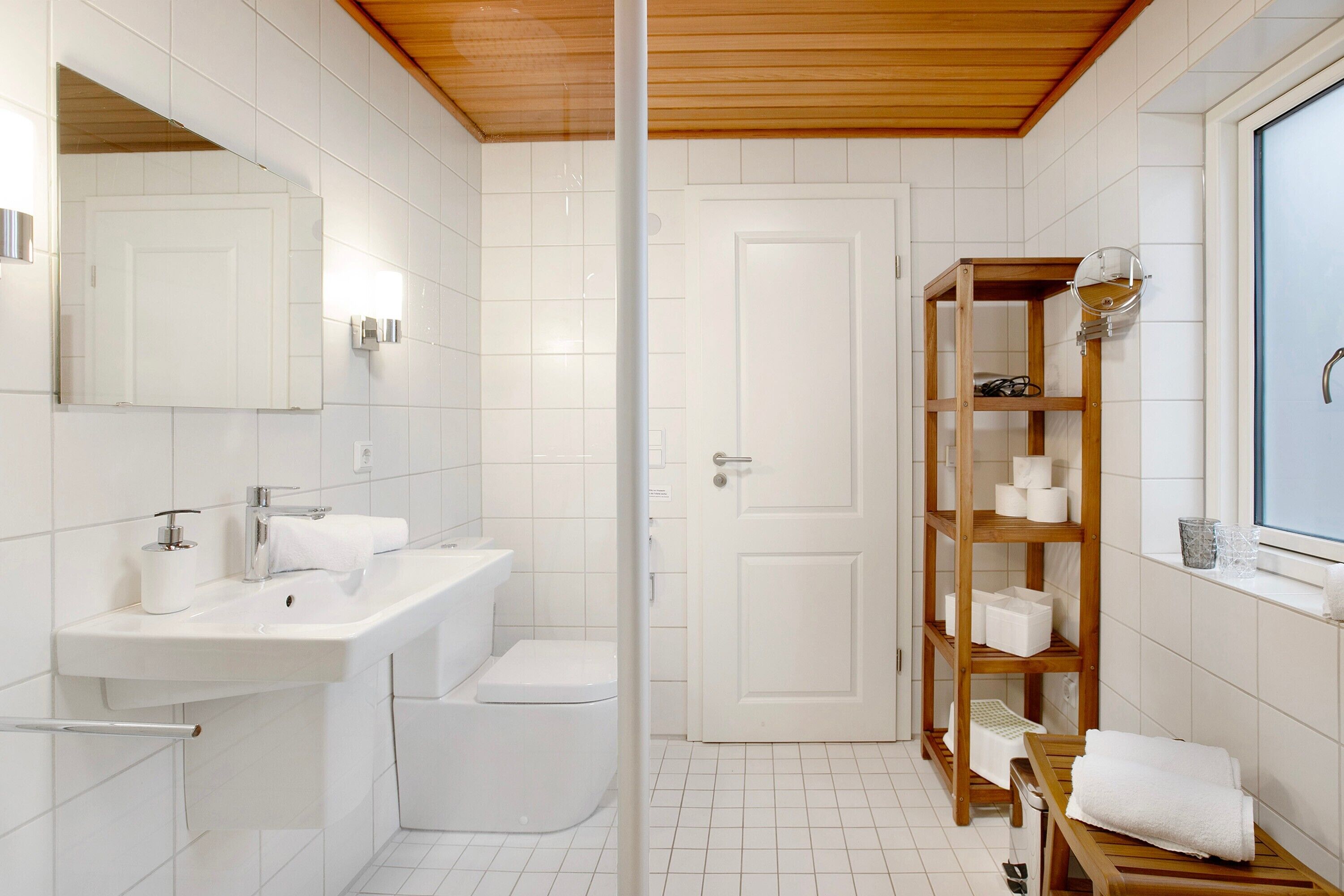 A modern bathroom with a white sink, toilet, and shower, wooden shelves with toiletries, and a large mirror.