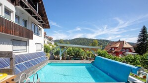 Una piscina con bordo in legno, pannelli solari e vista su una collina boscosa.