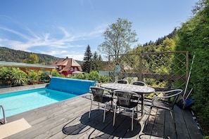 Eine Holzterrasse mit Pool, Gartenmöbeln und Blick auf einen bewaldeten Hang.