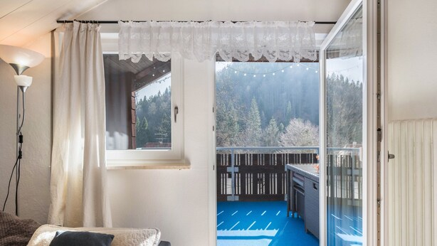 Una stanza con un'ampia porta scorrevole in vetro che si apre su un balcone con un tappetino blu, una lampada bianca e un divano con cuscini.