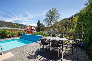 Eine Holzterrasse mit Pool, Gartenmöbeln und Blick auf einen bewaldeten Hang.