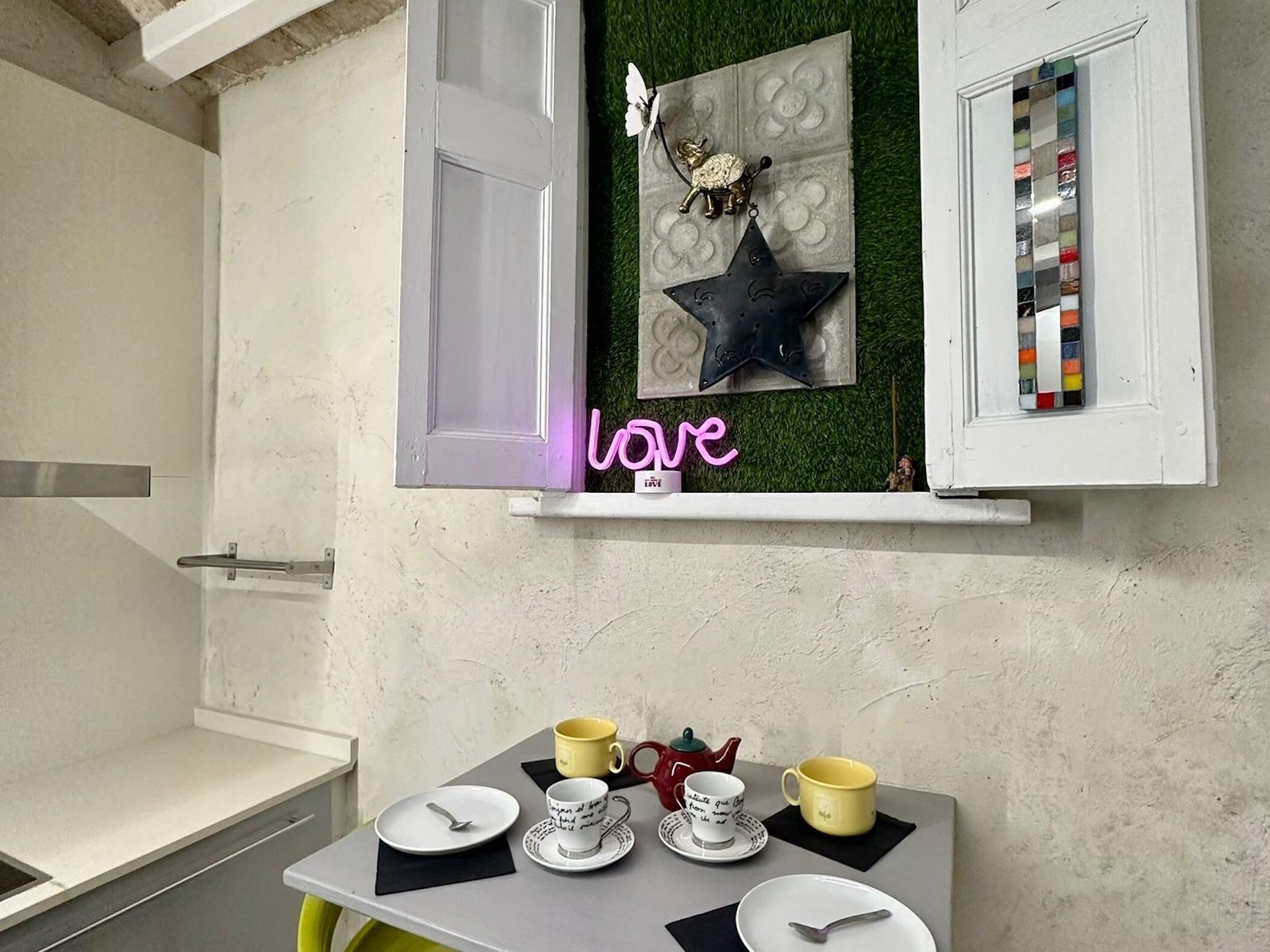 Une pièce comprenant un coin repas avec une table mise, sur laquelle on trouve des tasses, une théière et des assiettes. Au-dessus de la table, il y a une étagère décorative sur laquelle est accroché un panneau néon avec le mot « amour ».