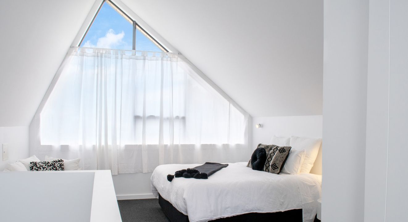 Un dormitorio con techo inclinado, una cama con ropa de cama blanca y una ventana con cortinas traslúcidas.