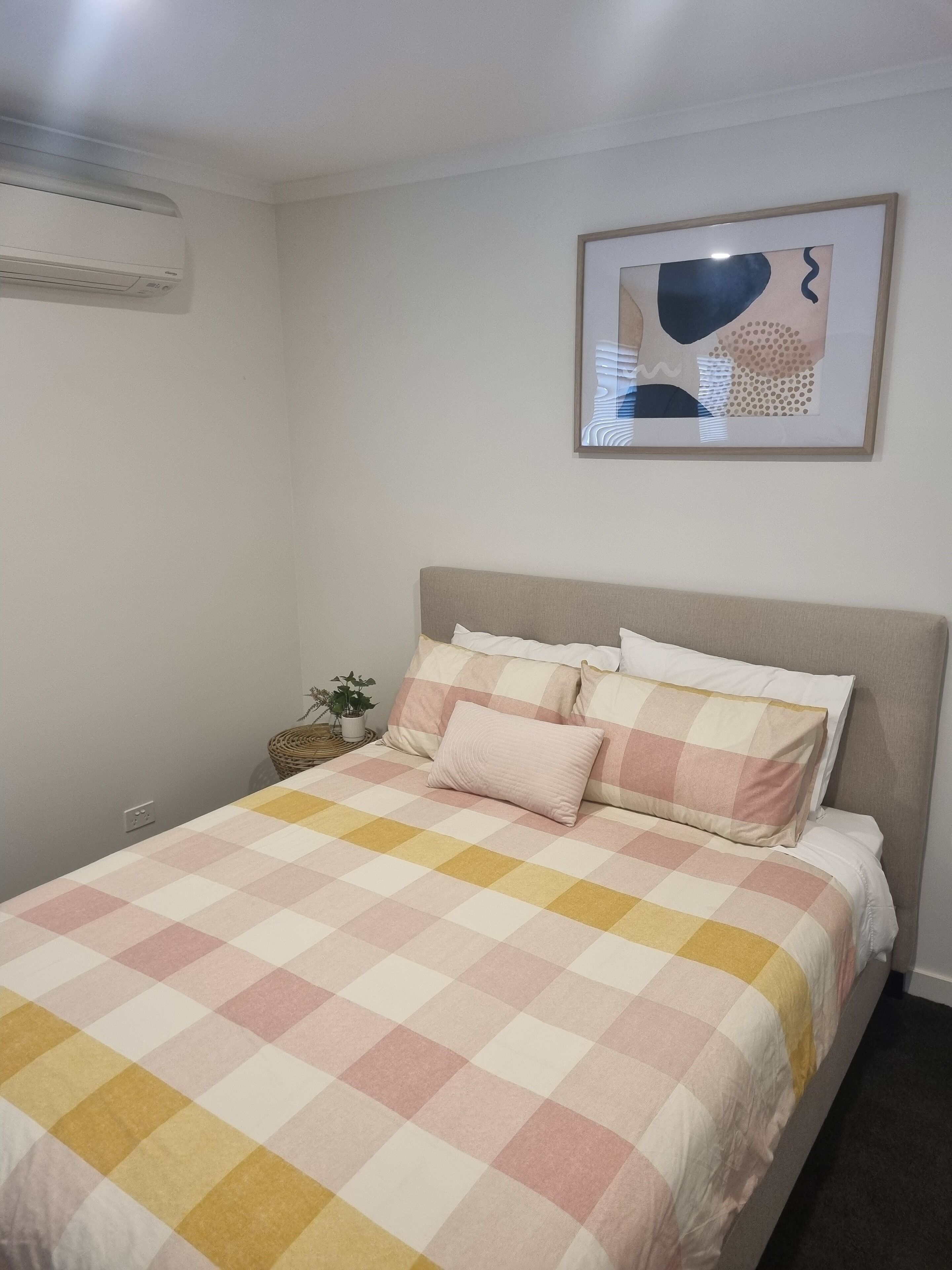 A bedroom with a checkered bedspread, a headboard, a wall-mounted air conditioner, a framed abstract painting, and a small potted plant.