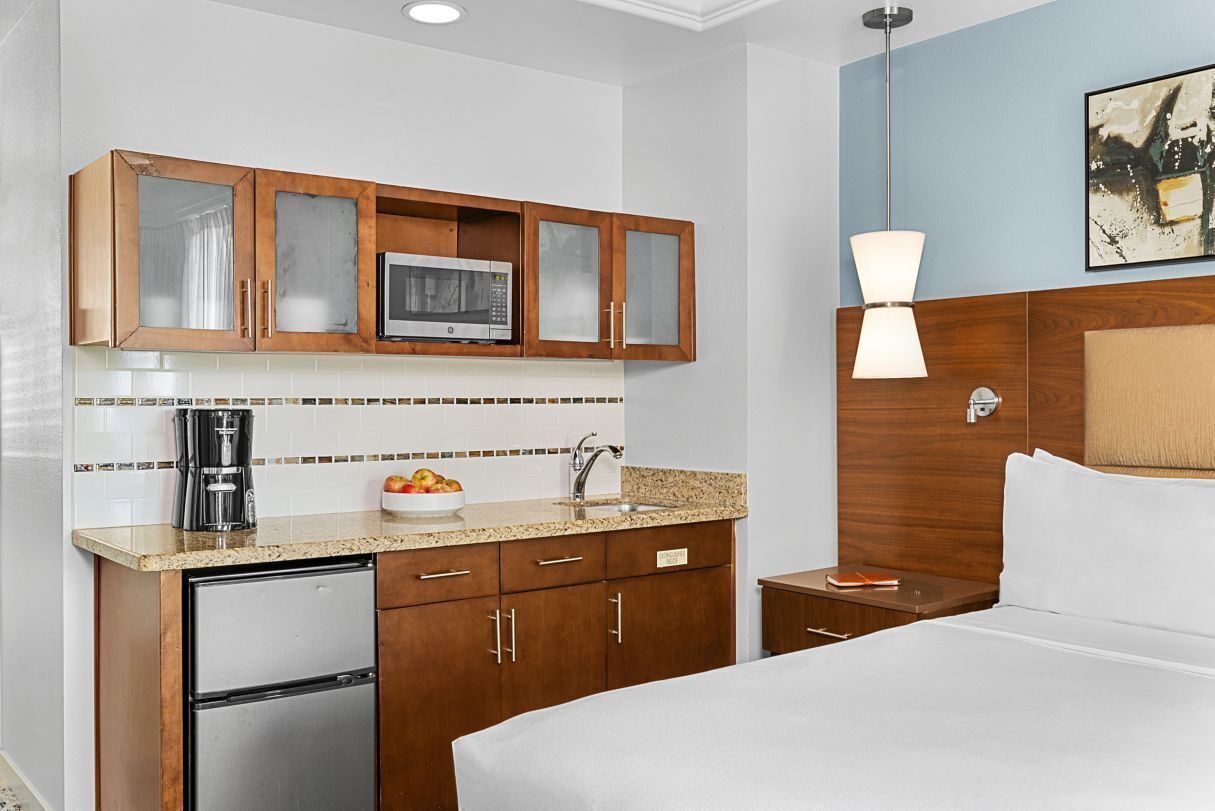 A compact hotel room with a kitchenette, a bed, a bedside table, and a nightstand.