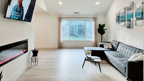 A modern living room with a flat-screen TV, a ceiling fan, a large window with curtains, a grey sofa, a coffee table, and decorative wall art.