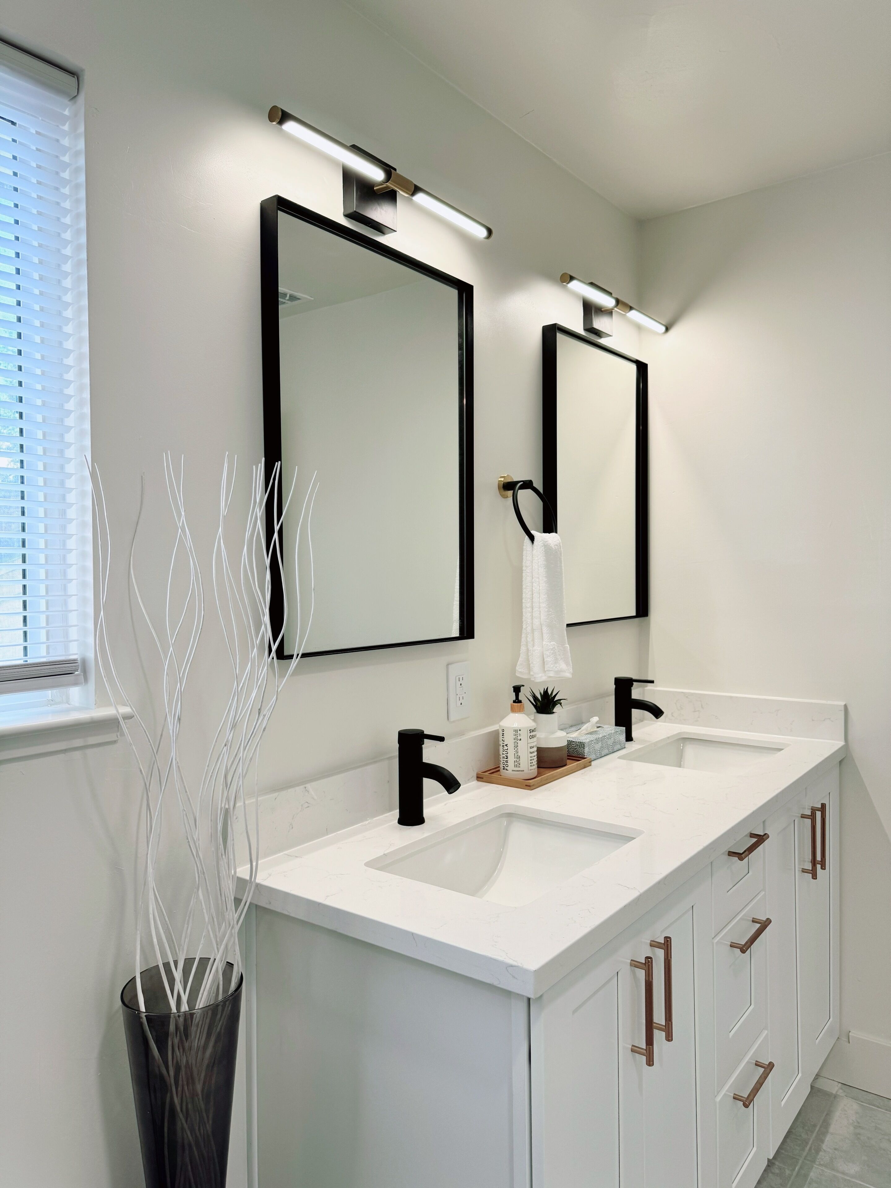 Een moderne badkamer met twee wasbakken, zwarte kraanwerk en een grote spiegel. Op het aanrecht staat een vaas met witte takken, en links een raam met luxaflex.