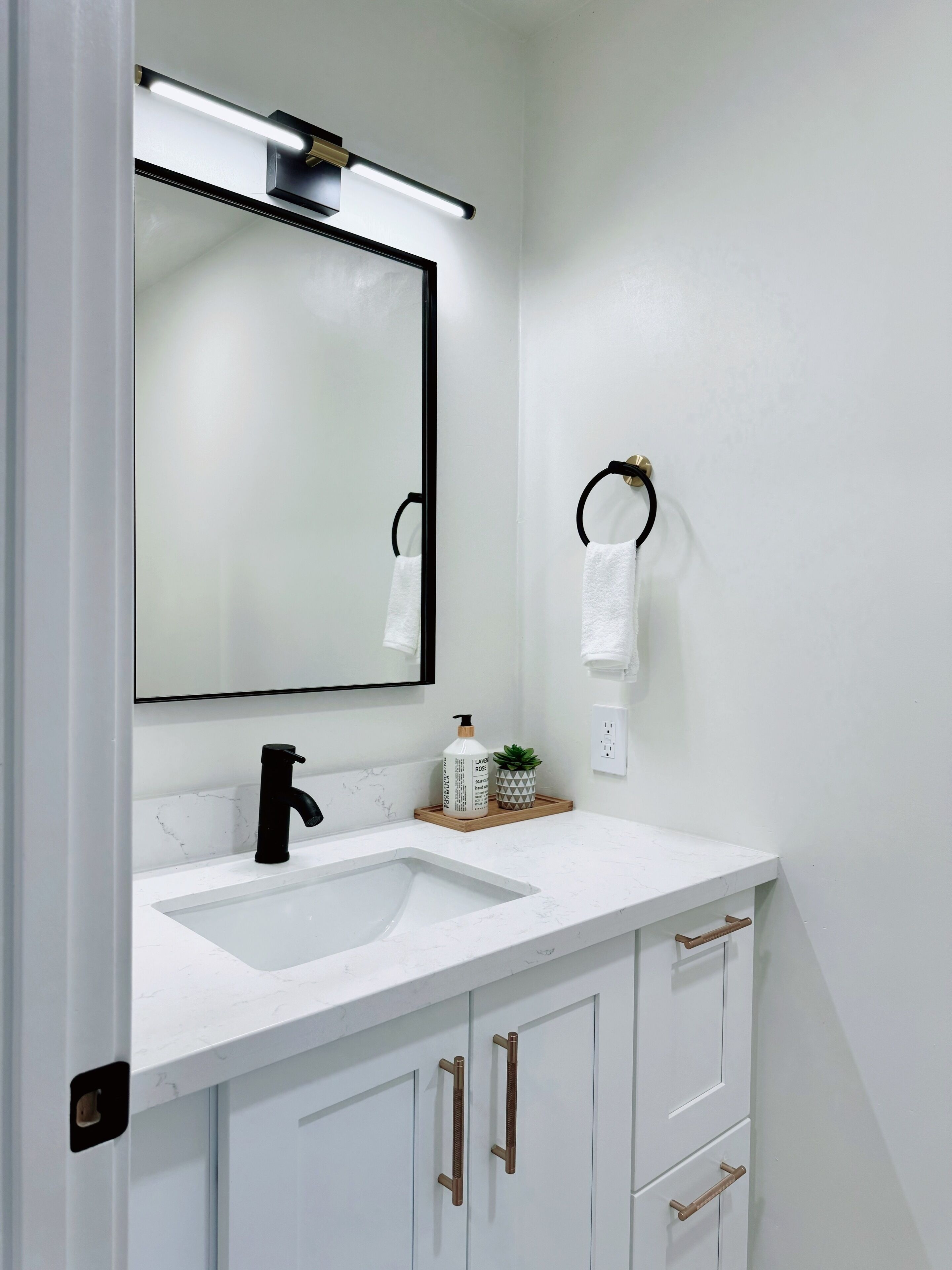 A modern bathroom with a white sink, black faucet, and a large mirror. There is a towel rack with a white towel, a small potted plant, and a bottle of hand soap on the counter.