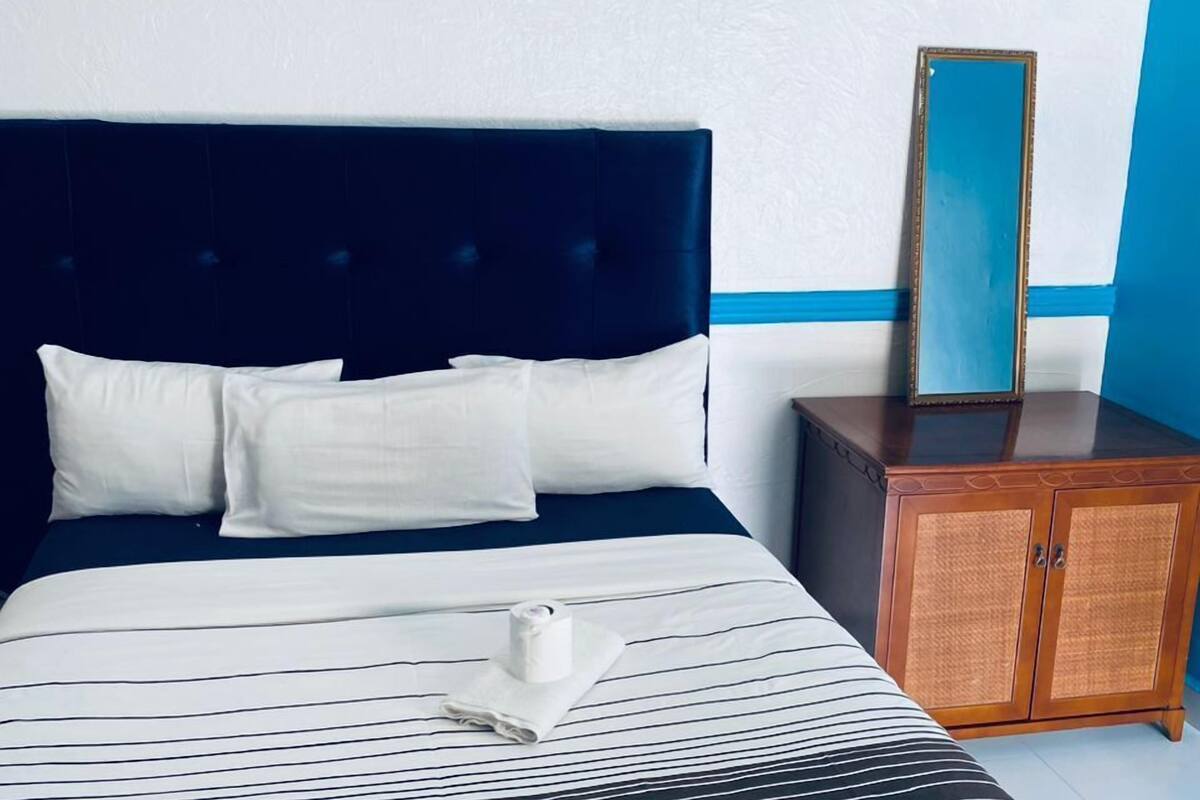 A neatly made bed with a striped bedspread, a dark headboard, and white pillows. There is a wooden nightstand with a mirror and a small cabinet.