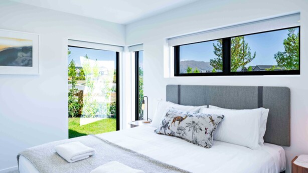 A modern bedroom with a large bed, a grey headboard, and a view of a garden through a sliding door.
