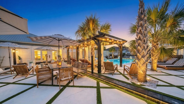 Zona de piscina con mobiliario de madera y una pérgola con luces colgantes.