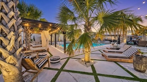 Un área junto a la piscina con sillones de descanso, una pérgola con luces y una palmera.