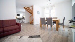 A modern living room with a red sofa, a dining table with four chairs, a staircase, and a kitchen in the background.