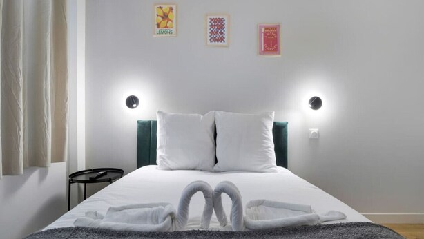 A neatly made bed with white linens and two pillows, a small bedside table, and wall-mounted lights.