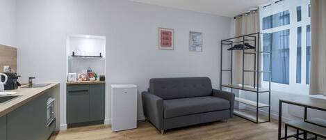 Un espacio habitable pequeño con kitchenette, sofá gris y ventana con cortinas.