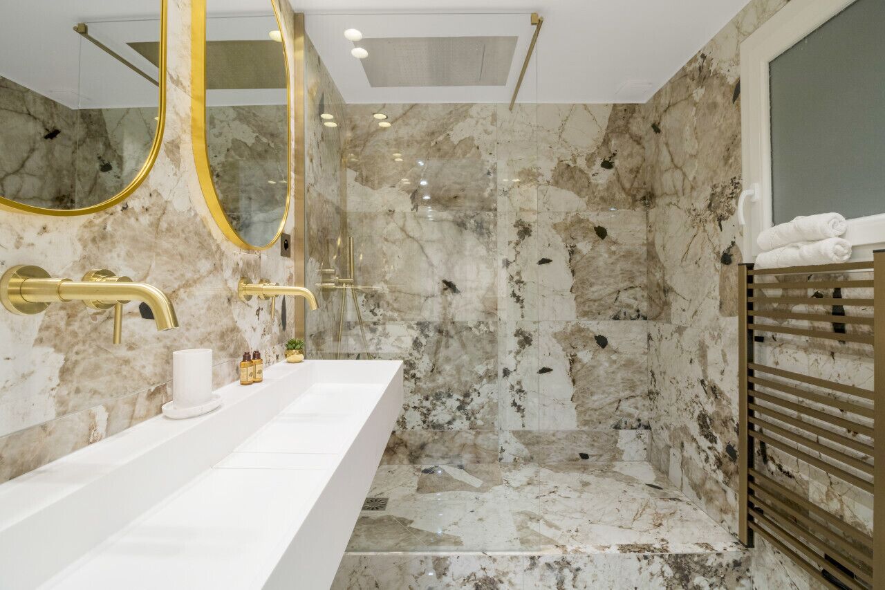 A modern bathroom with marble walls, a large mirror, gold fixtures, and a walk-in shower.