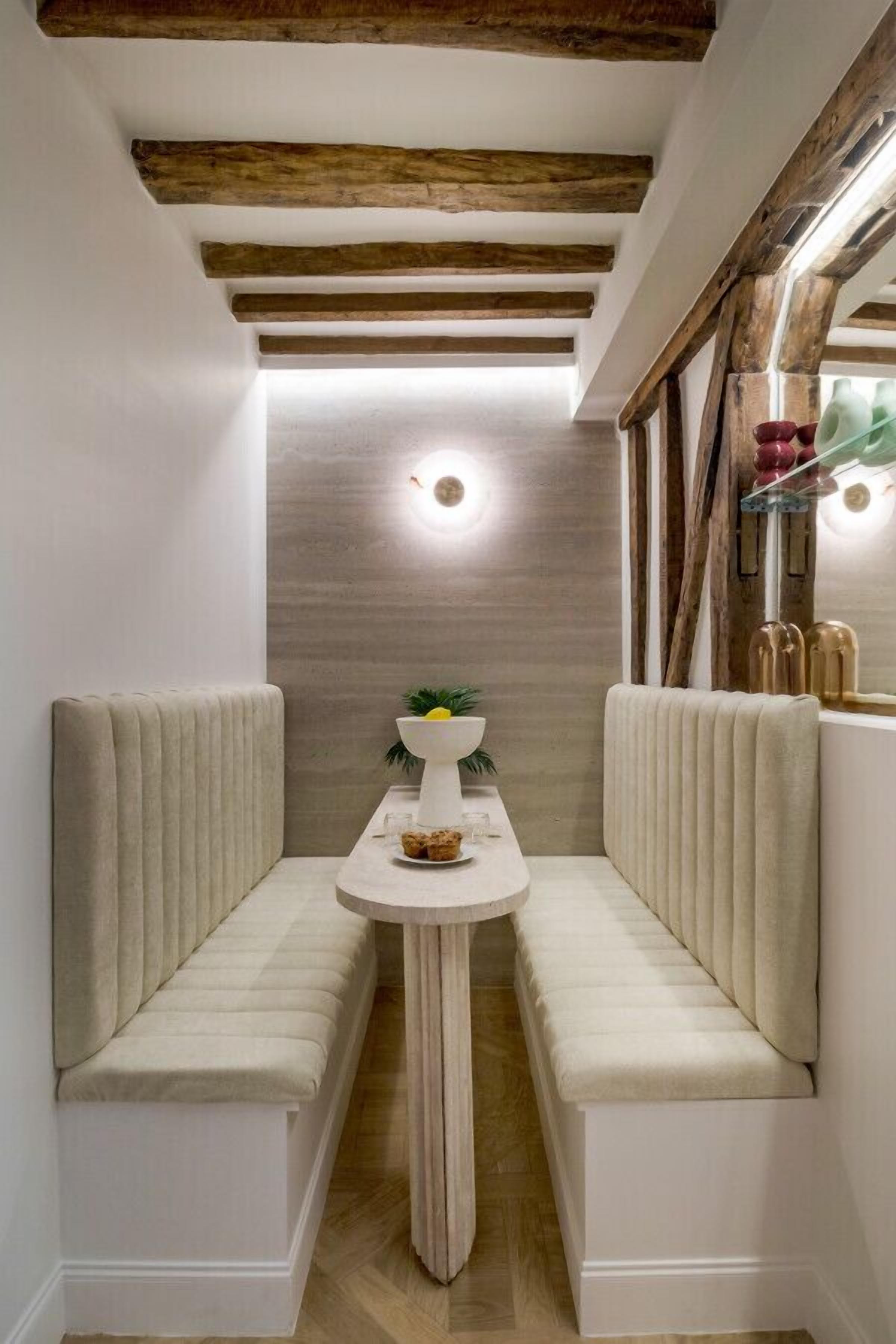 Un couloir étroit avec un banc servant de coin salon, une table ronde avec un saladier et des verres, et un plafond en bois avec des poutres apparentes.