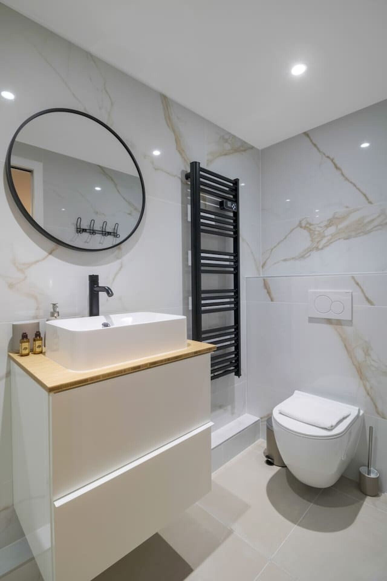 Une salle de bain moderne avec un pare-feu en marbre, un lavabo sur pied, un porte-serviettes noir, des toilettes et un miroir rond.