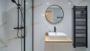 Un baño moderno con ducha envuelta en vidrio, un lavamanos blanco con grifo negro, un espejo redondo y un toallero negro.