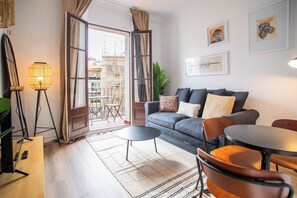A living room with a sofa, a coffee table, a dining table, and a floor lamp. There is a balcony with a view of buildings. - 43APAR1010 - Super Centric Apartment in Barcelona (Barcelona)