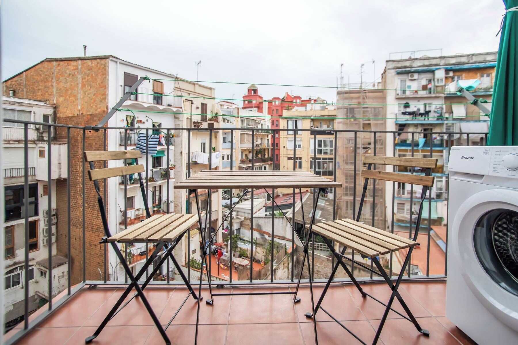 A balcony with a table and chairs, a washing machine, and a view of a residential area with multi-story buildings.
