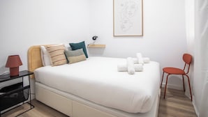 Ein ordentlich eingerichtetes Schlafzimmer mit Bett, Kissen, einem Nachttisch, einem Sessel, einem gerahmten Kunstwerk und einem Regal mit Lampe.