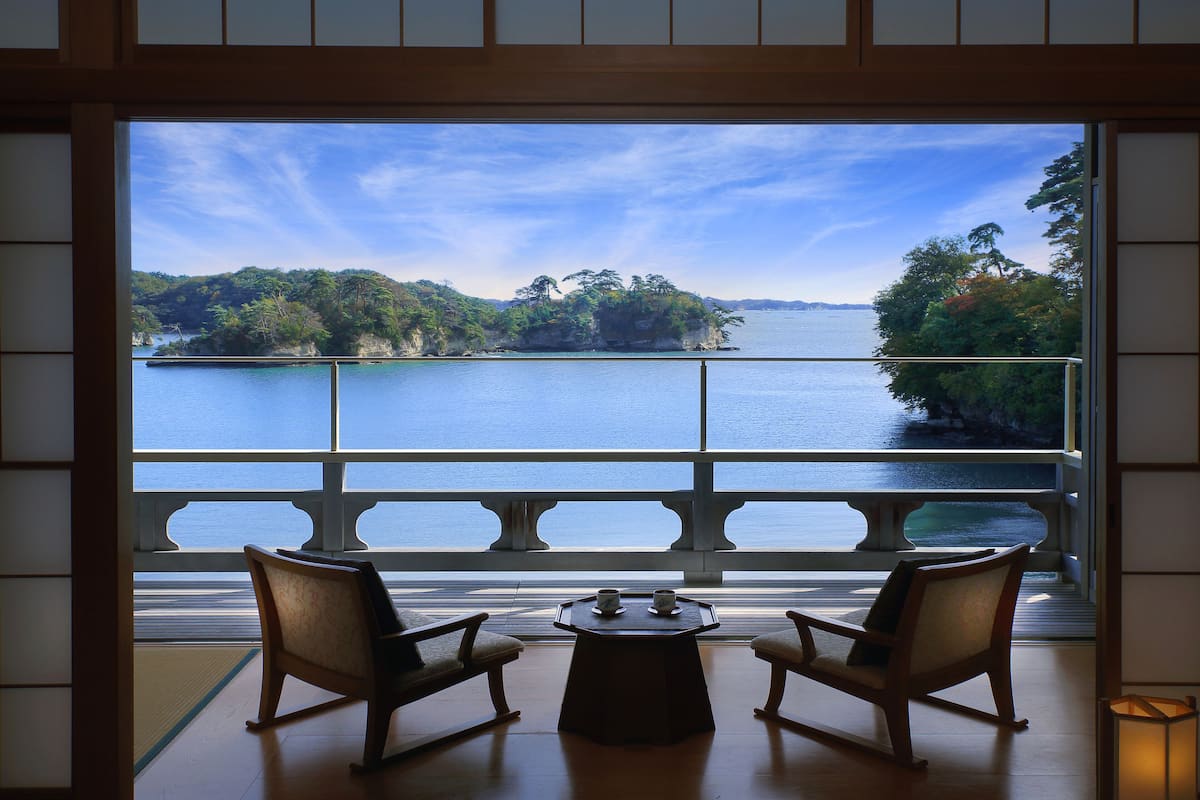 A room with a view of a lake and mountains, two chairs, and a small table with cups.