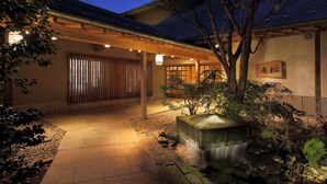 A traditional Japanese building with a stone pathway leading to a wooden gate, surrounded by greenery and a small waterfall feature.