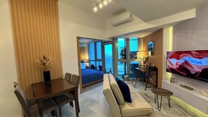 Una sala de estar moderna con sofá, mesa de comedor y sillas. Hay un televisor en la pared y un ventanal con vistas a la ciudad.