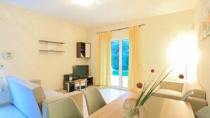 A modern living room with a sofa, a dining table, and a TV. There is a sliding glass door with curtains. - 3 Bedroom Villa, Private Pool, Agios Stefanos (Agios Stefanos)