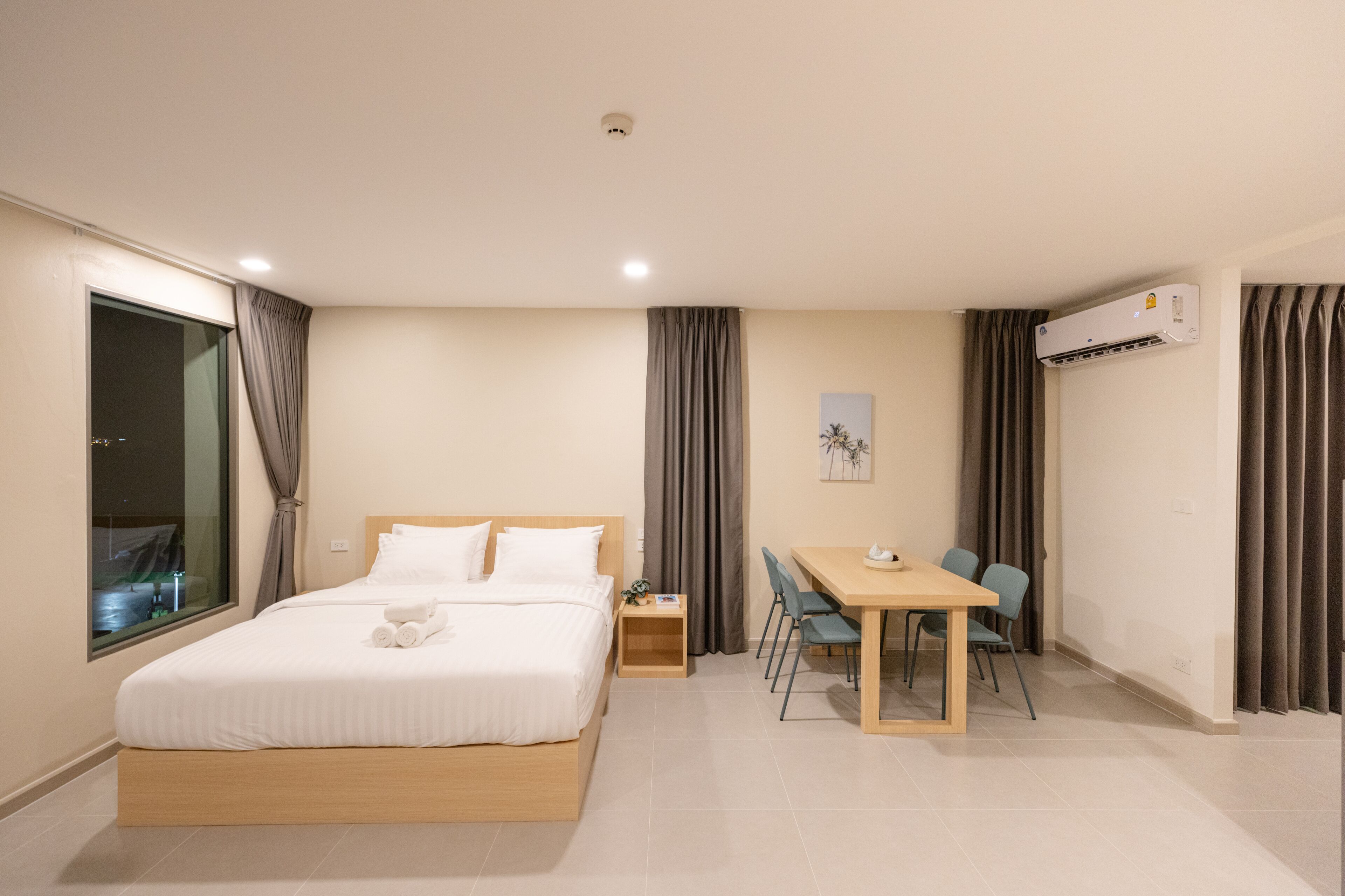 A hotel room with a bed, bedside table, desk, chairs, and an air conditioning unit.