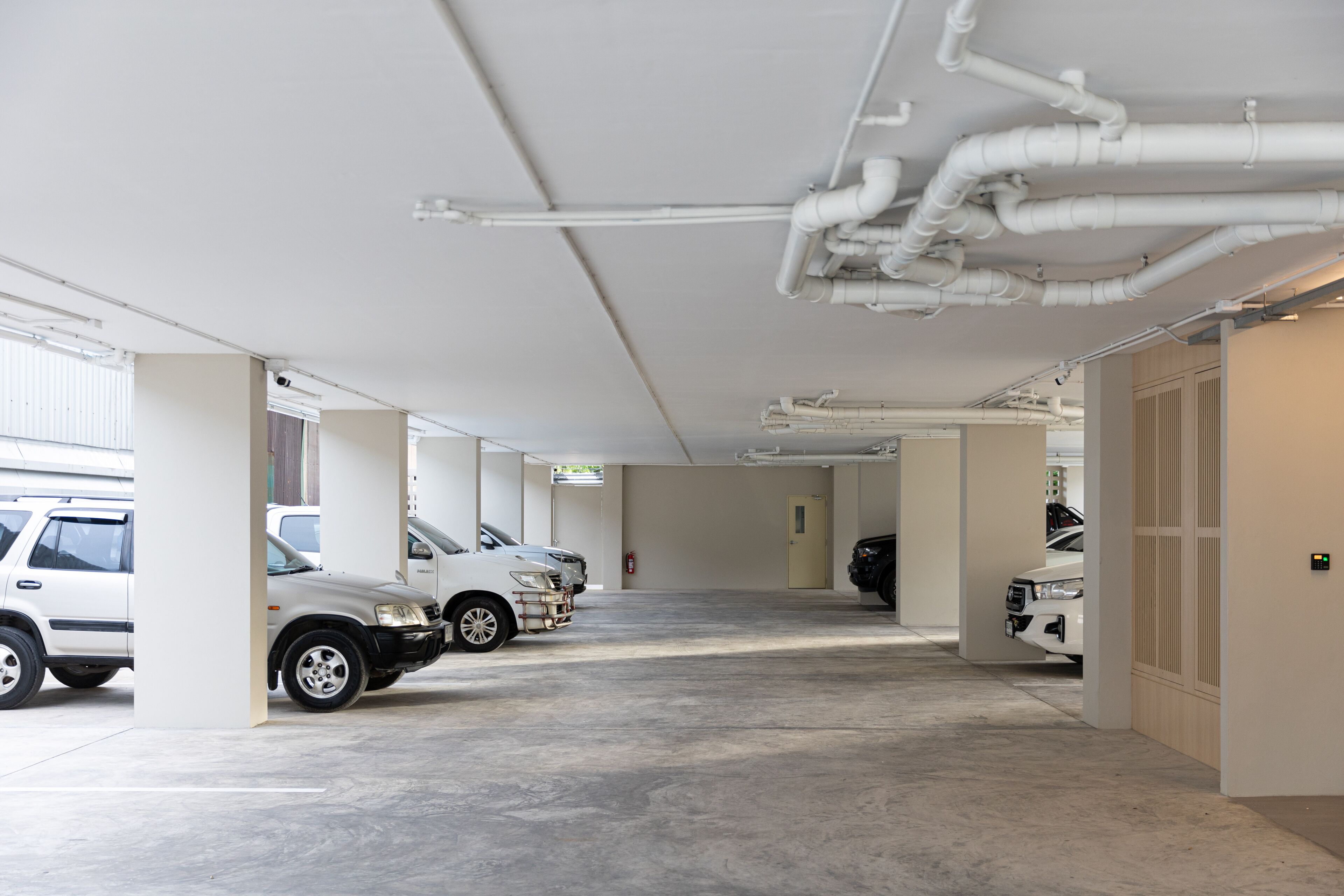 Un parcheggio multipiano con diverse auto parcheggiate, pareti bianche e tubature a vista sul soffitto.