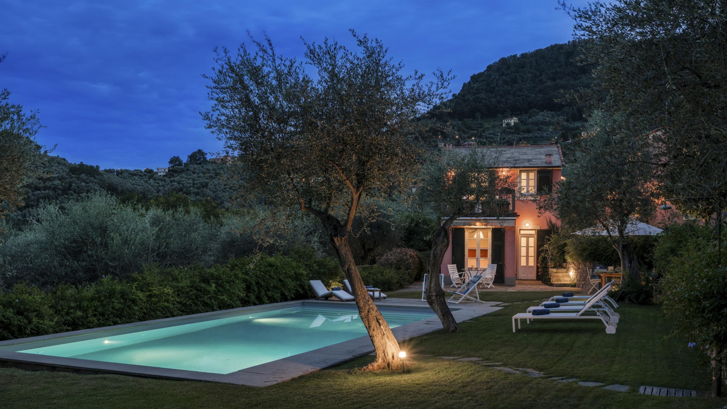 A poolside area with lounge chairs and a swimming pool, surrounded by lush greenery and a hillside in the background.