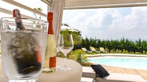 A glass of water, a bottle of champagne, and a wine glass on a table by a pool with lounge chairs and greenery in the background. - Villa Lisa 8 Emma Villas (Rimini)