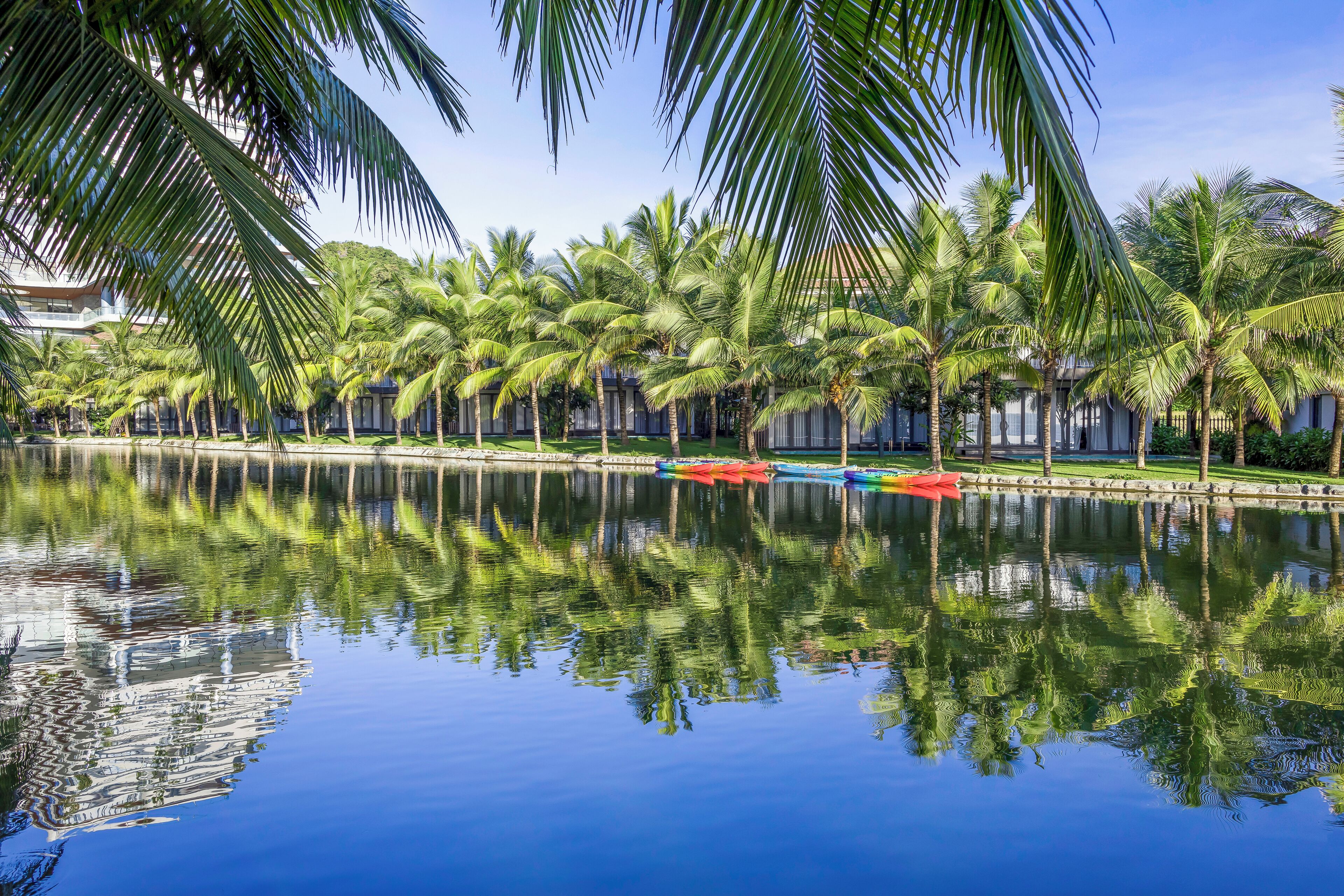 A tranquil water body reflecting palm trees and a building with a balcony.
