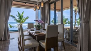 Uma sala de jantar com vista para o oceano, uma mesa posta com pratos e um vaso de flores, e uma varanda com uma palmeira.