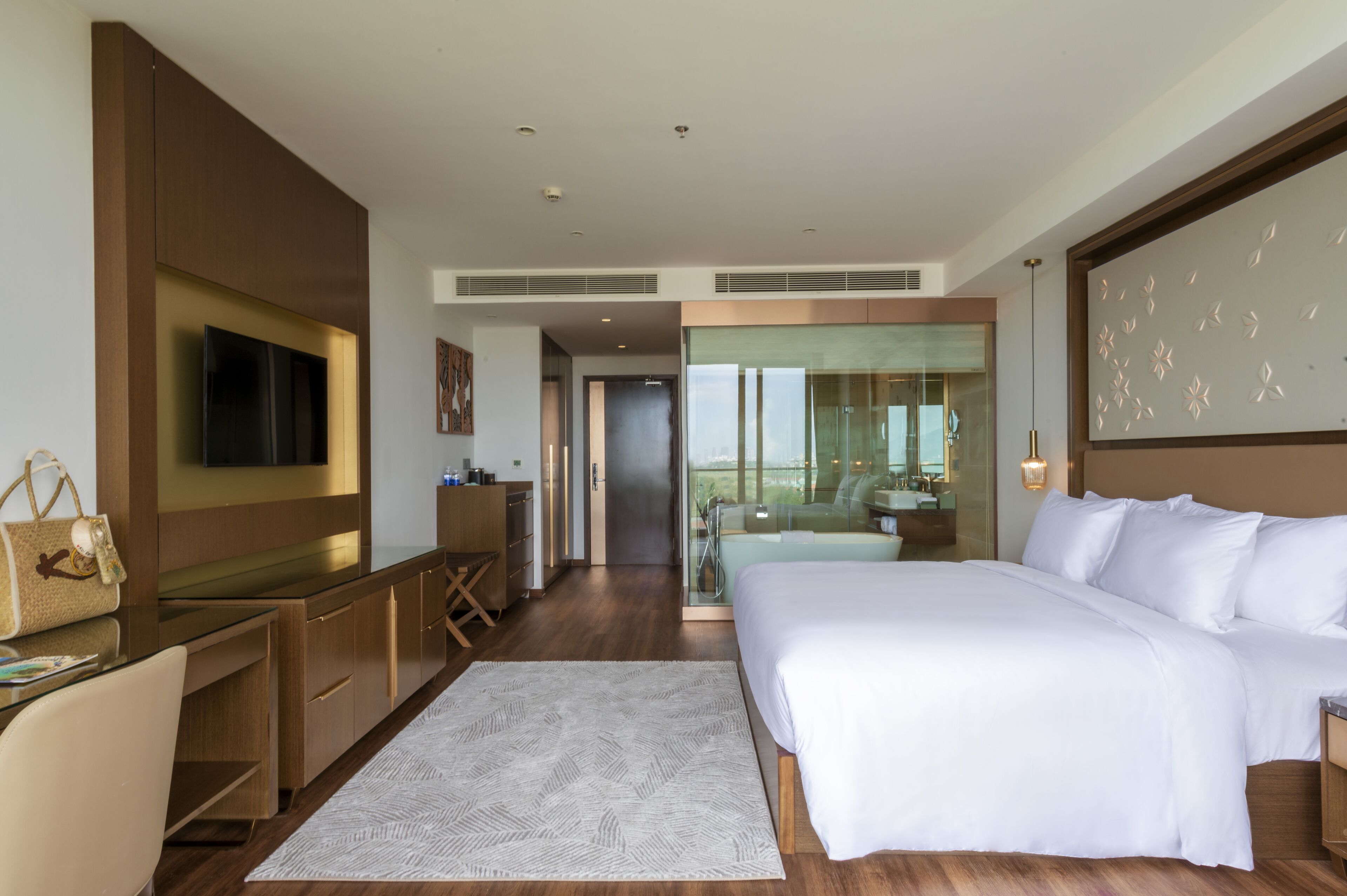 A modern hotel room with a large bed, a desk with a flat-screen TV, a glass-enclosed bathroom, and a view of the city.