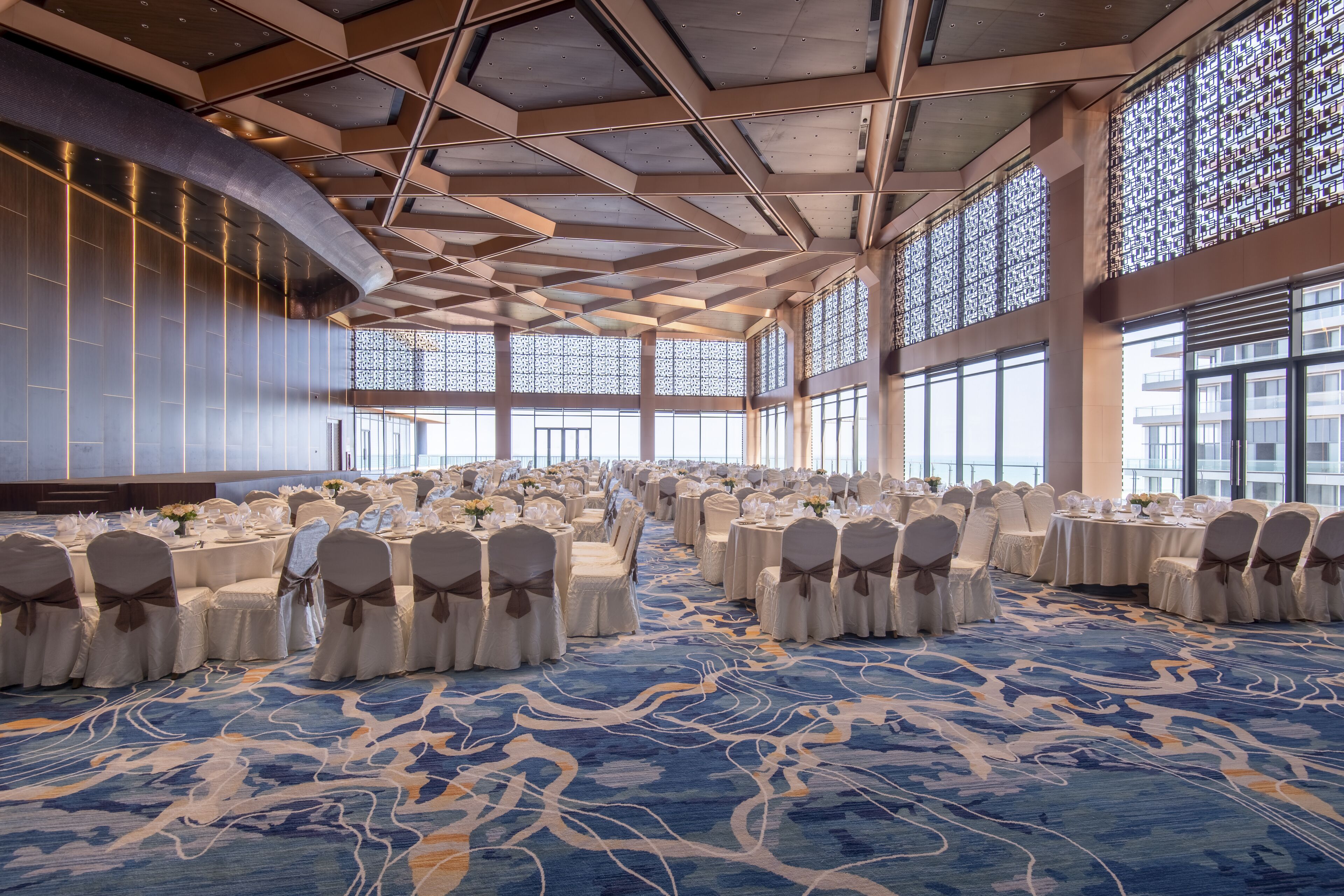 Un salón de banquetes con mesas redondas, sillas blancas y alfombra azul.
