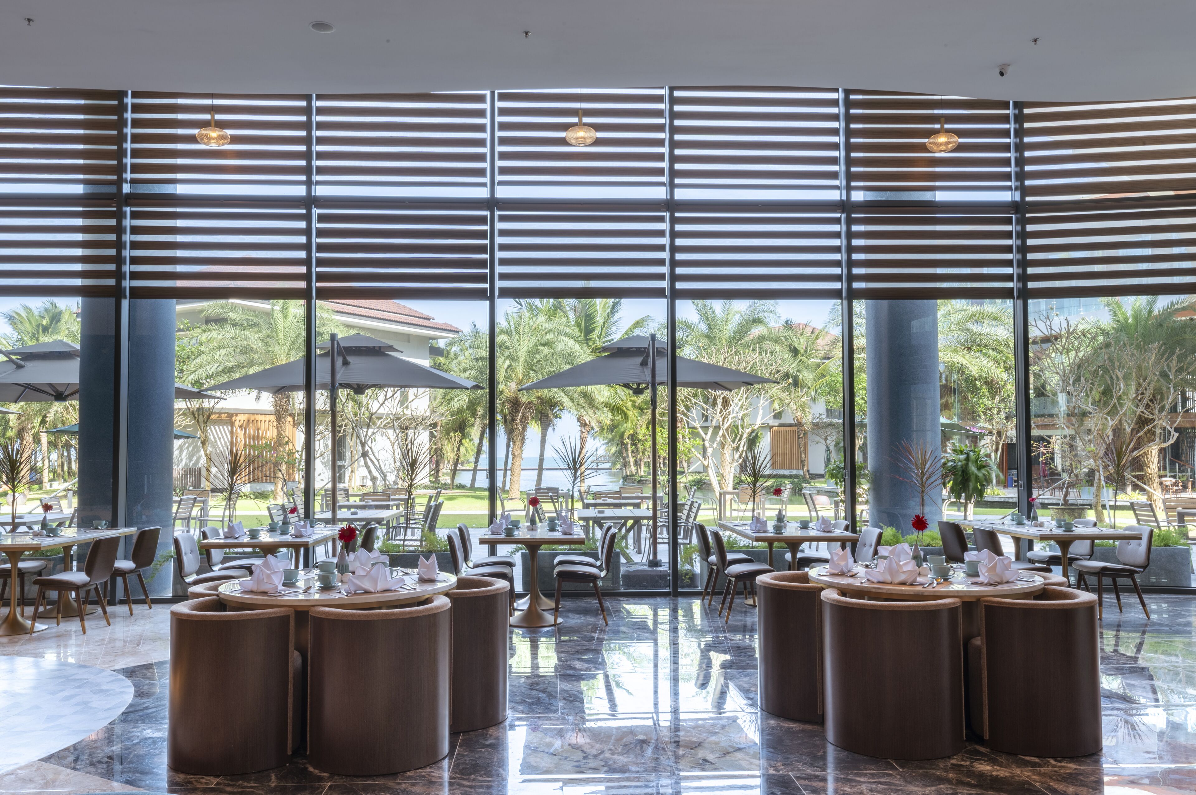 Un amplio espacio exterior para comer con mesas y sillas redondas, ventanas grandes con persianas y vistas a palmeras y un edificio al fondo.