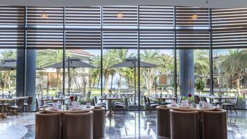 Un amplio espacio exterior para comer con mesas y sillas redondas, ventanas grandes con persianas y vistas a palmeras y un edificio al fondo.