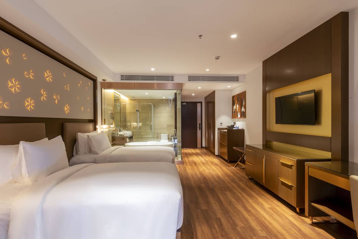 Une chambre d’hôtel avec deux lits, une télévision, un bureau et une salle de bain séparée par une paroi en verre.