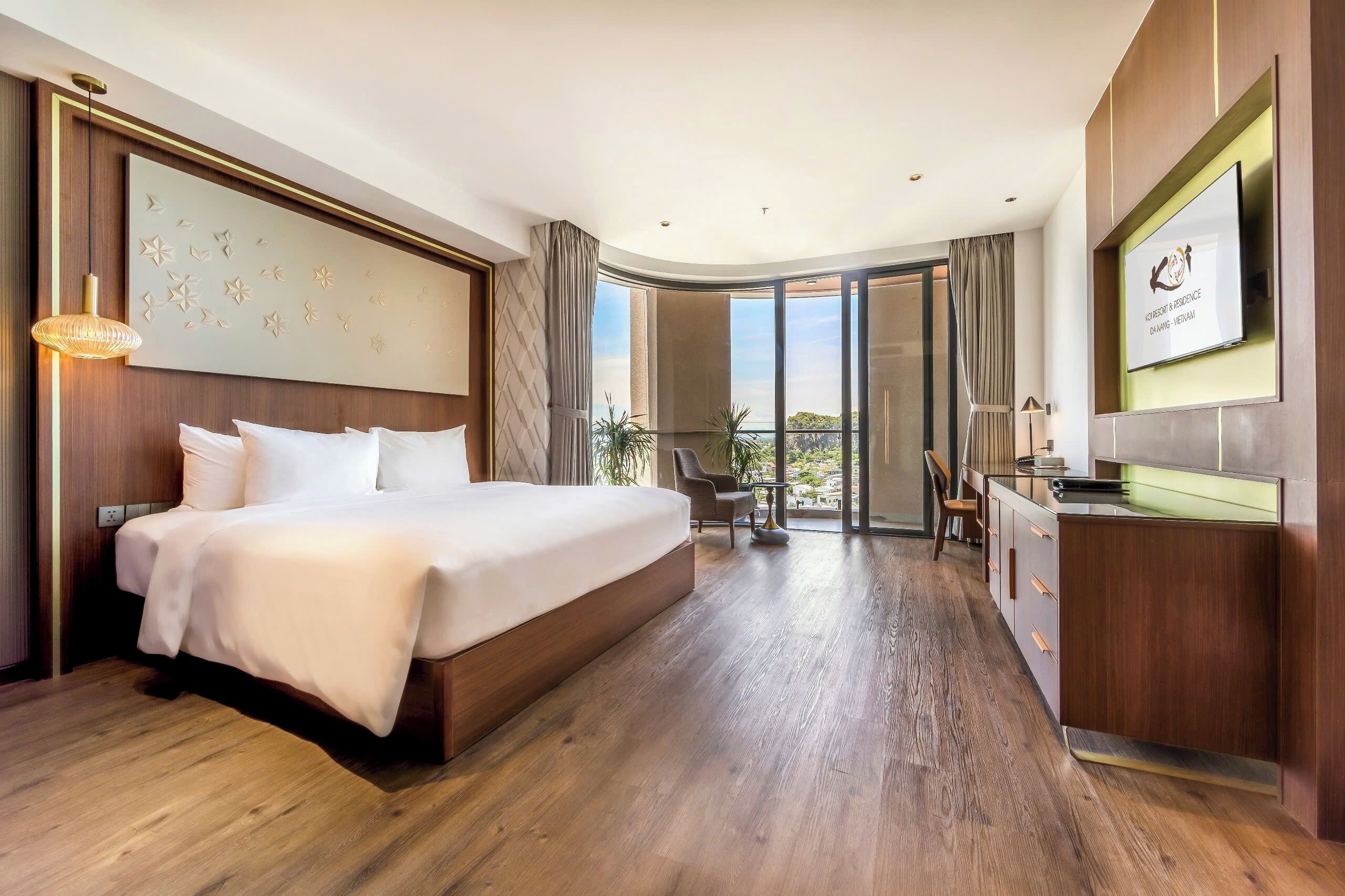 A hotel room with a large bed, a desk, a TV, and a balcony with a view.
