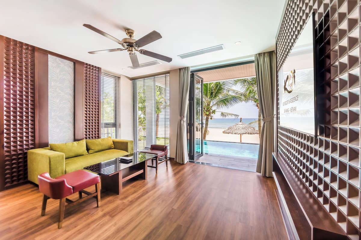 Une chambre d’hôtel moderne avec un plancher en bois, un canapé vert, un fauteuil rouge, un ventilateur de plafond et une vue sur la plage et l’océan, visible à travers la porte vitrée.