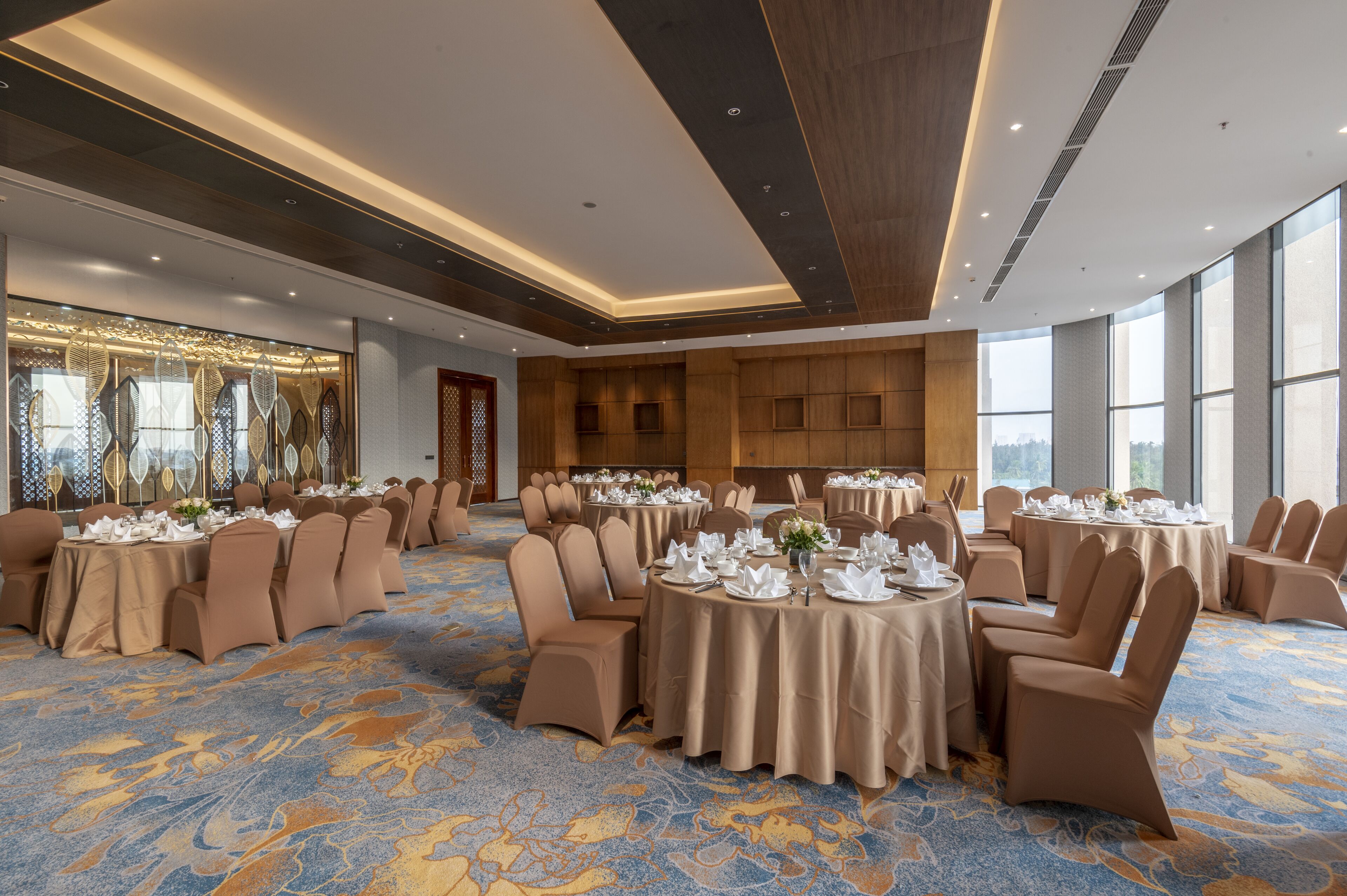 A banquet hall with round tables set for dining, large windows, and a patterned carpet.