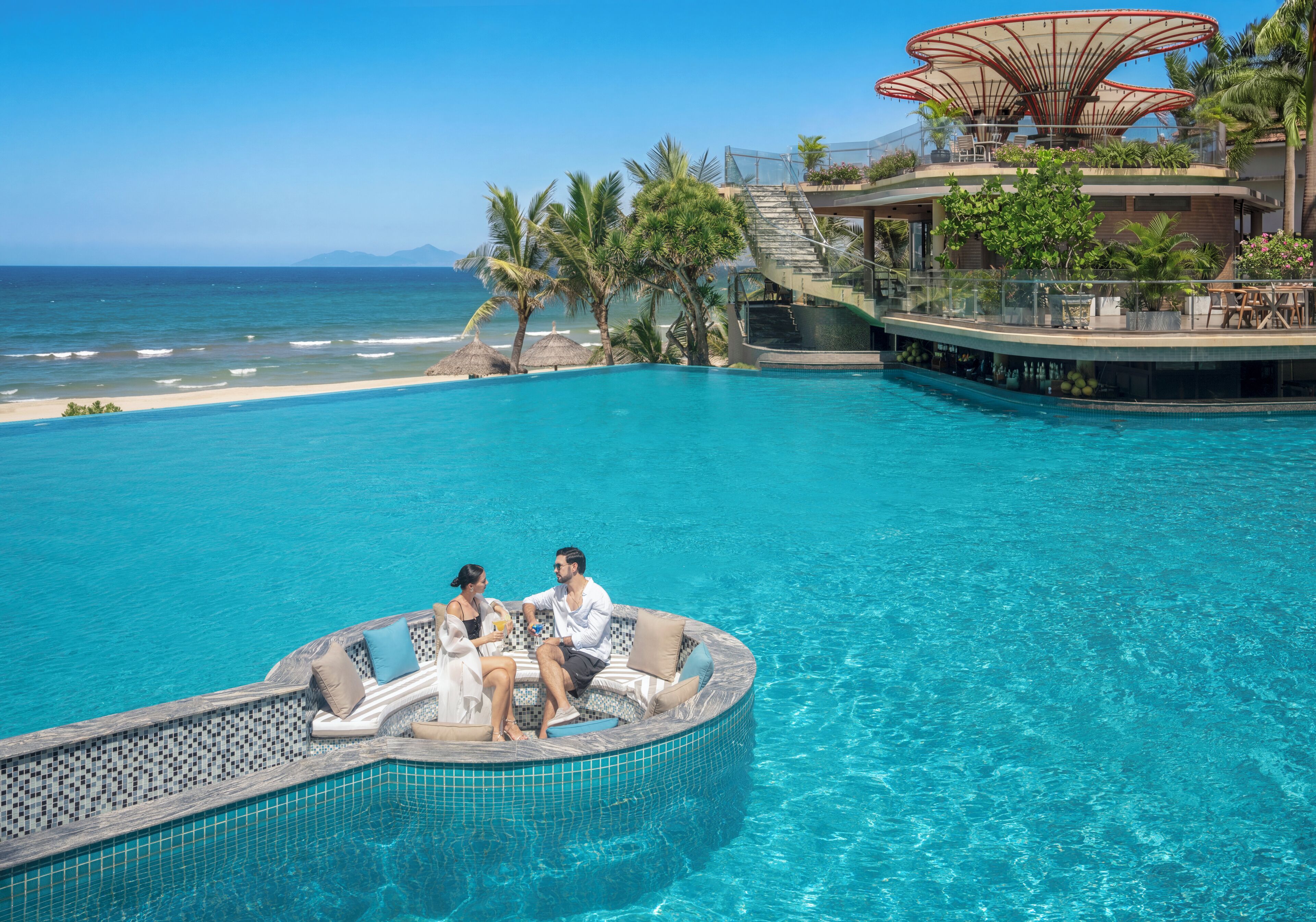 Una piscina con un área de descanso circular, una escalera que lleva a un edificio y vista a la playa.
