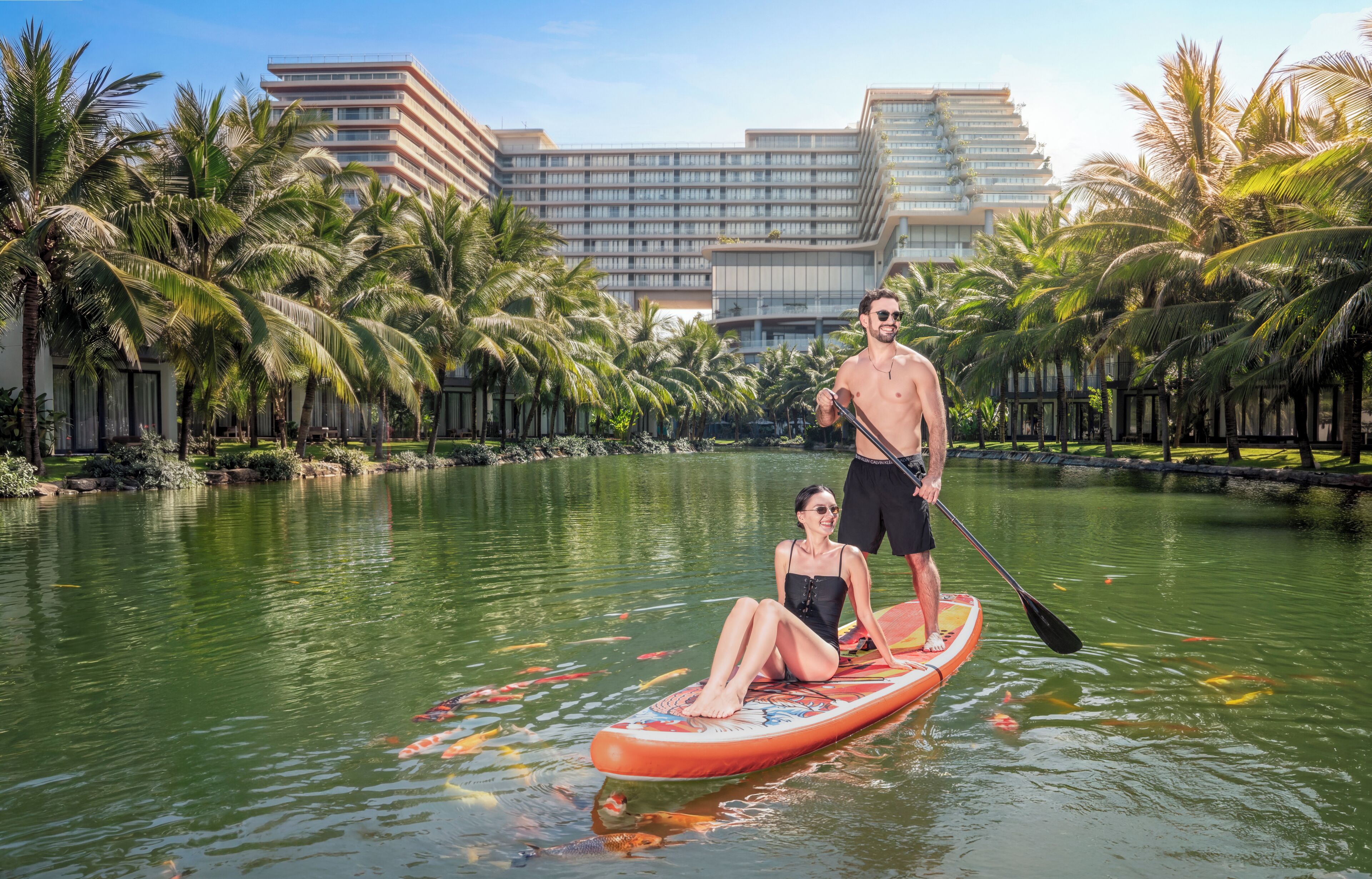 A couple on a paddleboard in a body of water with a hotel and palm trees in the background.
