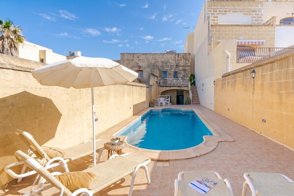 A swimming pool with lounge chairs and an umbrella, surrounded by stone walls and a building with a balcony.